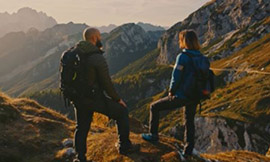 man and woman on a hike wearing backpacks and standing on top of a mountain while looking out at the sky